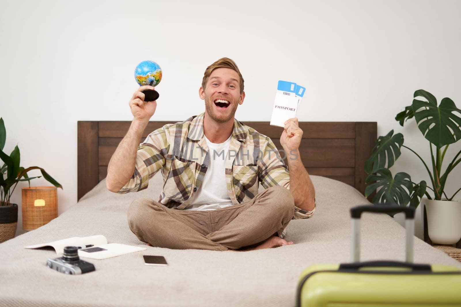 Portrait of happy guy, tourist goes on vacation abroad, holds globe and plane ticket, excited about his holiday, sits on bed with smiling, enthusiastic face expression. Copy space