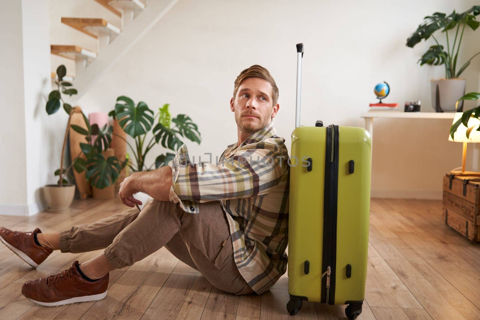 Portrait of male model with suitcase, sitting on floor, holding flight tickets and passport, going abroad, waiting for a taxi to airport.