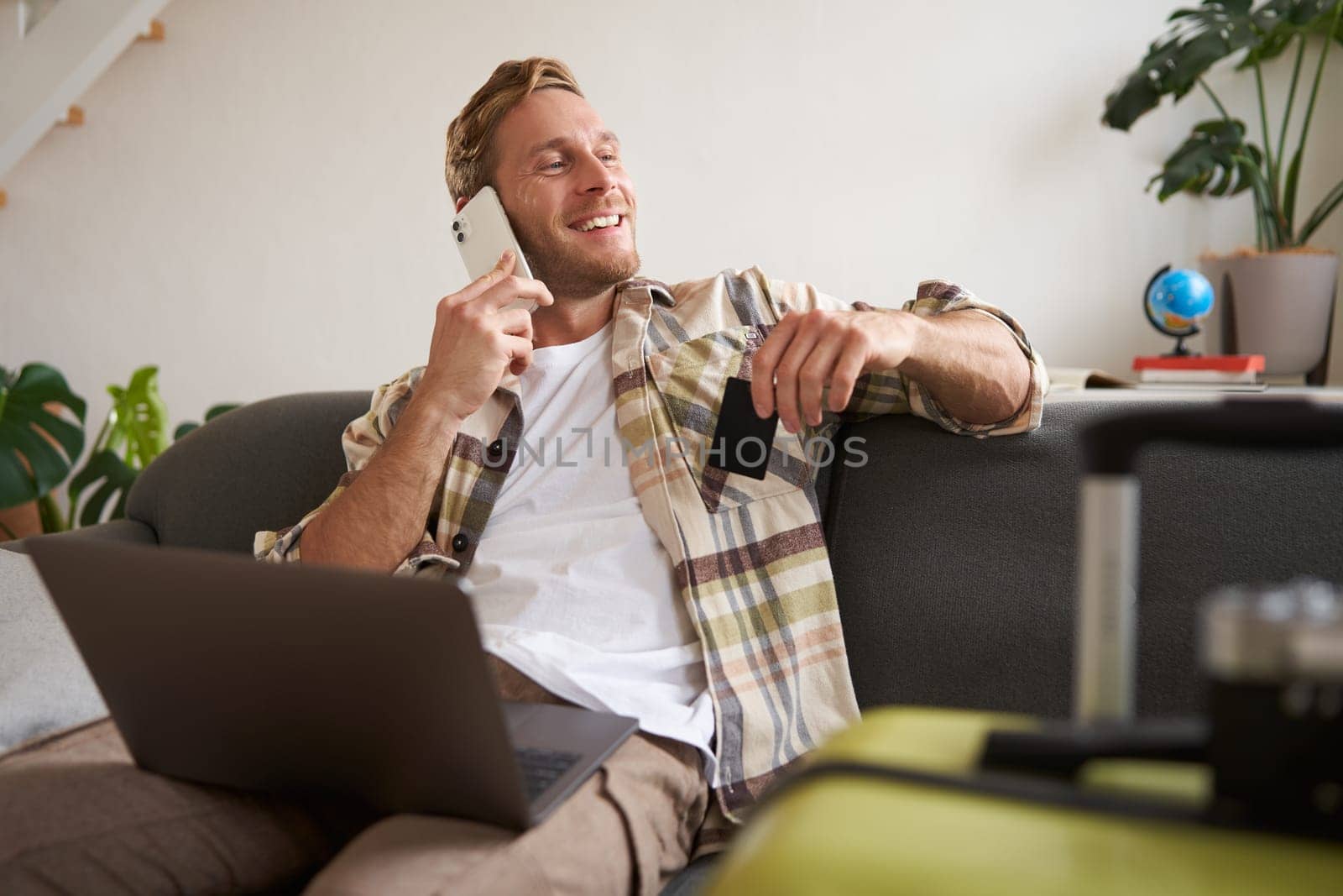 Smiling guy talking on mobile phone, sitting with laptop, holding credit card, looking out of the window with happy, relaxed face expression.