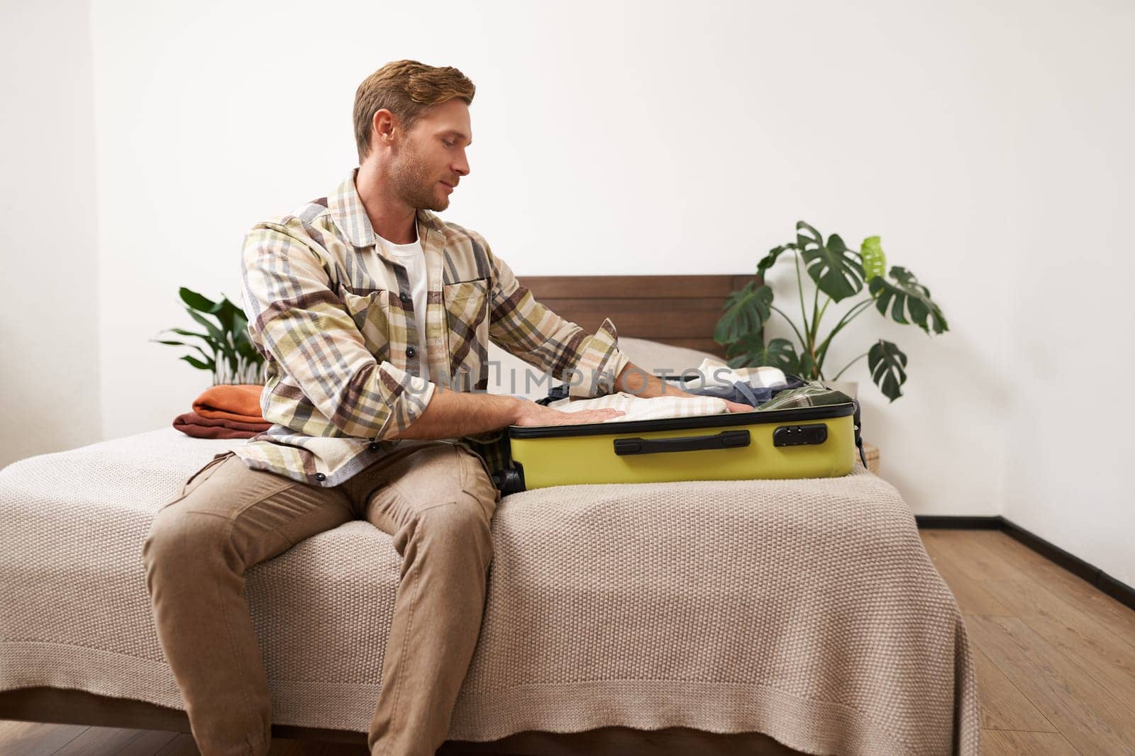 Portrait of young man packing his luggage for a trip, going on holiday, sitting on bed and putting clothes in suitcase, preparing for his flight.