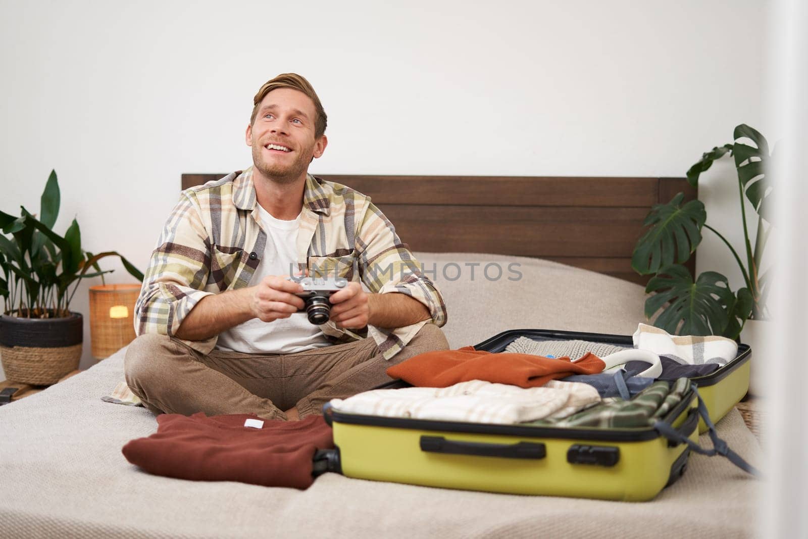 Portrait of young excited man, sits on bed with suitcase, going on holiday, holding digital camera, going through photo album on gadget, preparing for summer vacation.