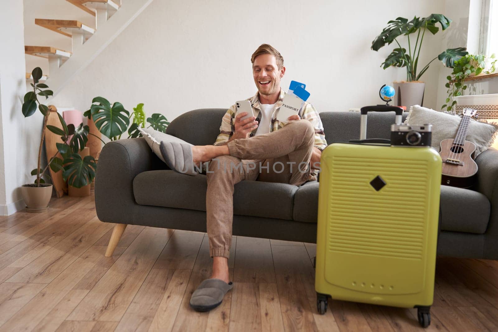 Portrait of man sits with mobile phone in living room, packed suitcase, chats on smartphone app, looks for holiday destinations, goes on vacation.