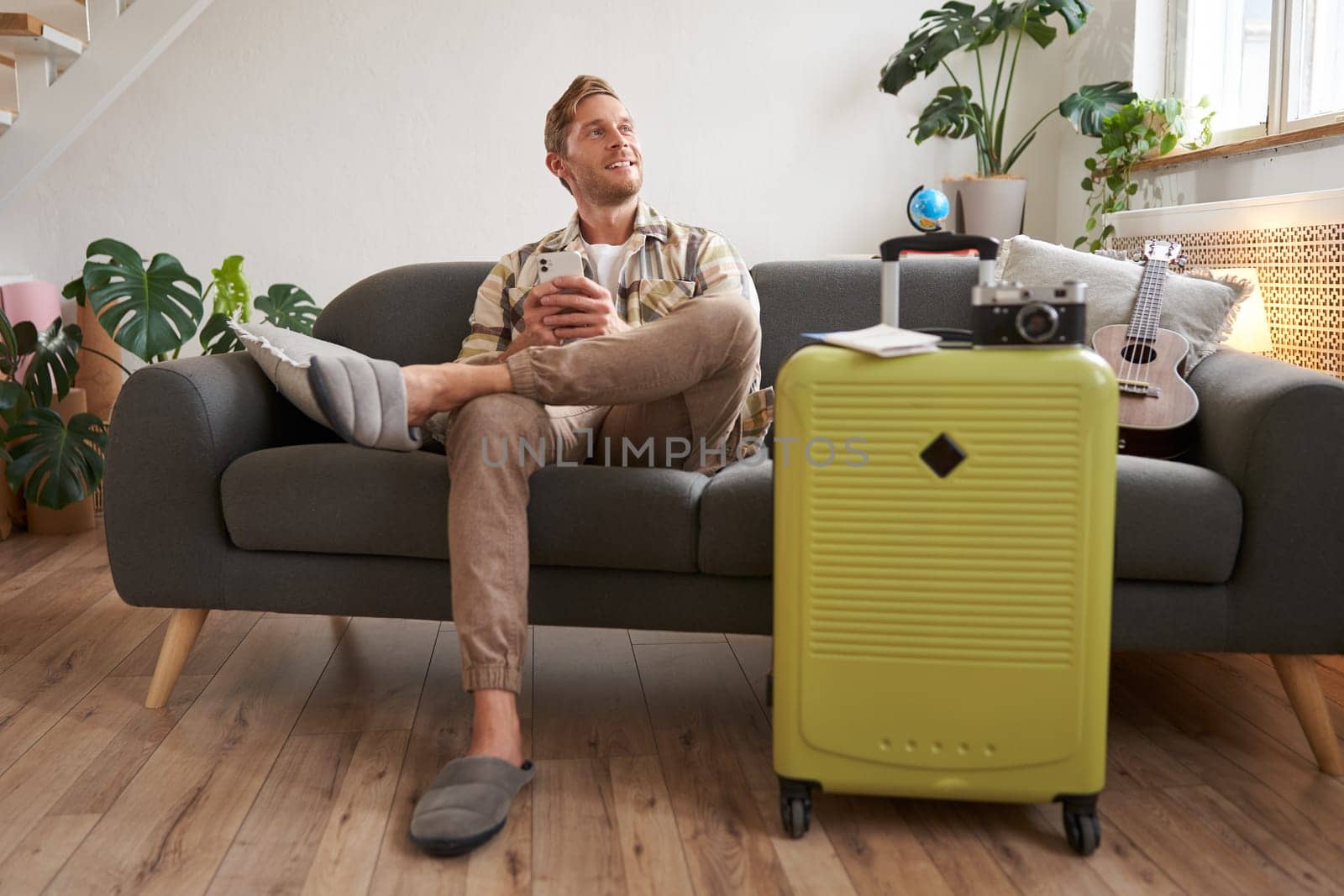 Portrait of young man sitting in living room, dreaming about holiday, sitting with suitcase and smartphone, looking aside thoughtful.