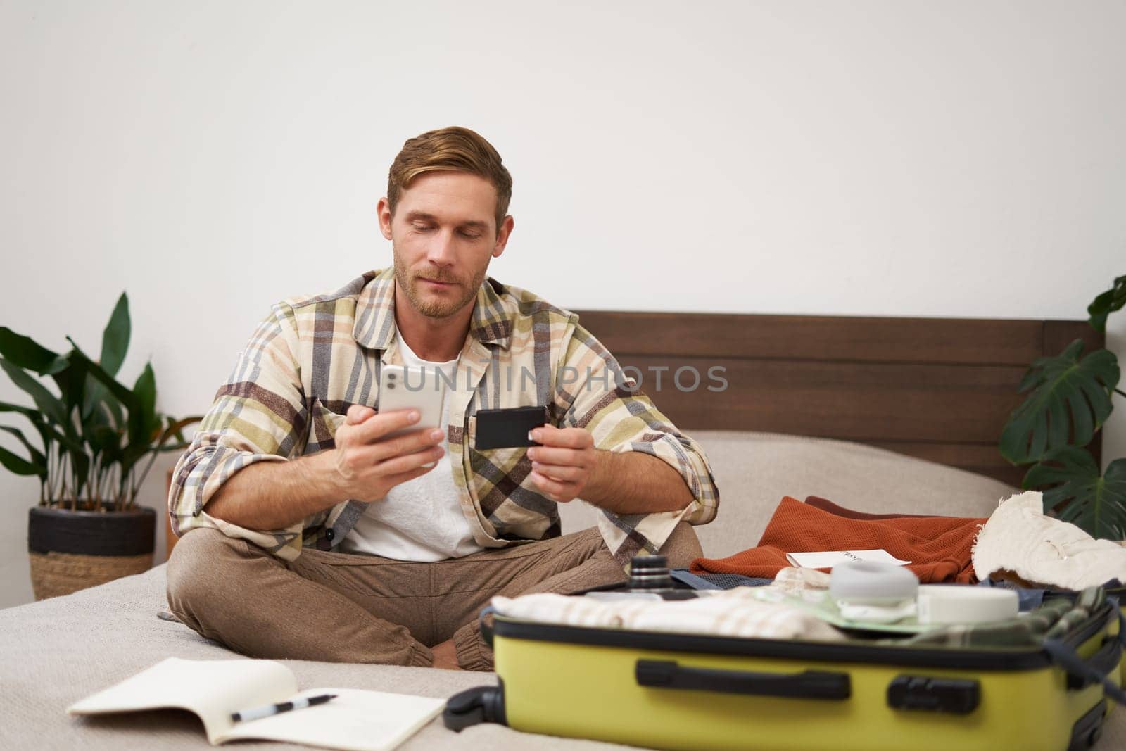 Portrait of happy man in suitcase, transferring money, buying tickets online, sitting on bed with credit card and smartphone app, paying online by Benzoix