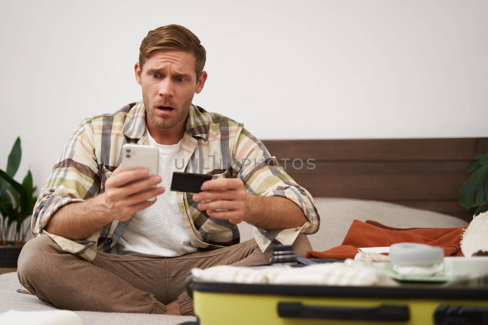 Portrait of worried young man, sitting on bed with suitcase, holding credit card and mobile phone, looking concerned at smartphone screen while packing for holiday. Transactions and finance concept