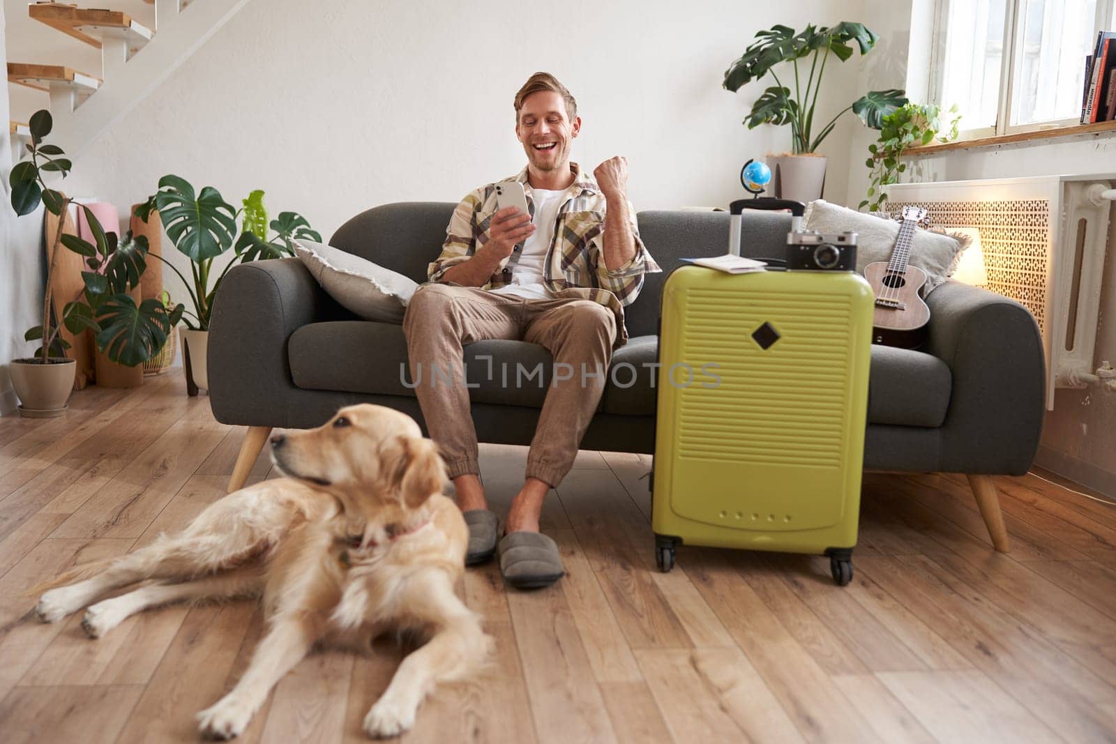 Happy man with his dog, sits in living room with suitcase, looks at smartphone app with excited face, pleased with booked trip by Benzoix