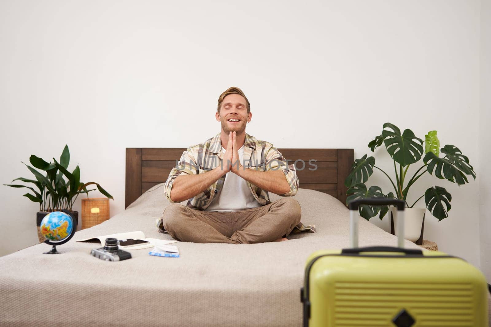 Portrait of traveller practice yoga, young man meditating before going on vacation, sitting with suitcase, globe and camera on bed in bedroom, relaxing, looking for peaceful holiday by Benzoix