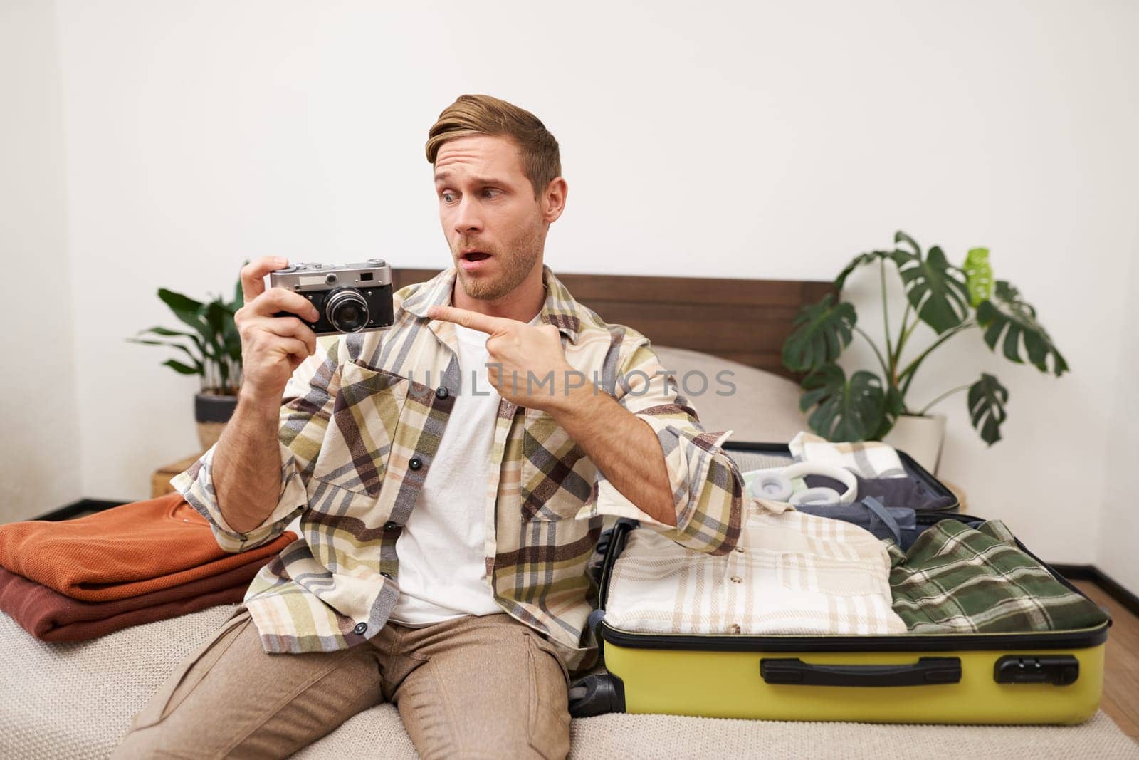 Portrait of handsome happy man, holding his analogue camera, sitting near opened suitcase, packing things for holiday, travelling abroad, getting ready for vacation.
