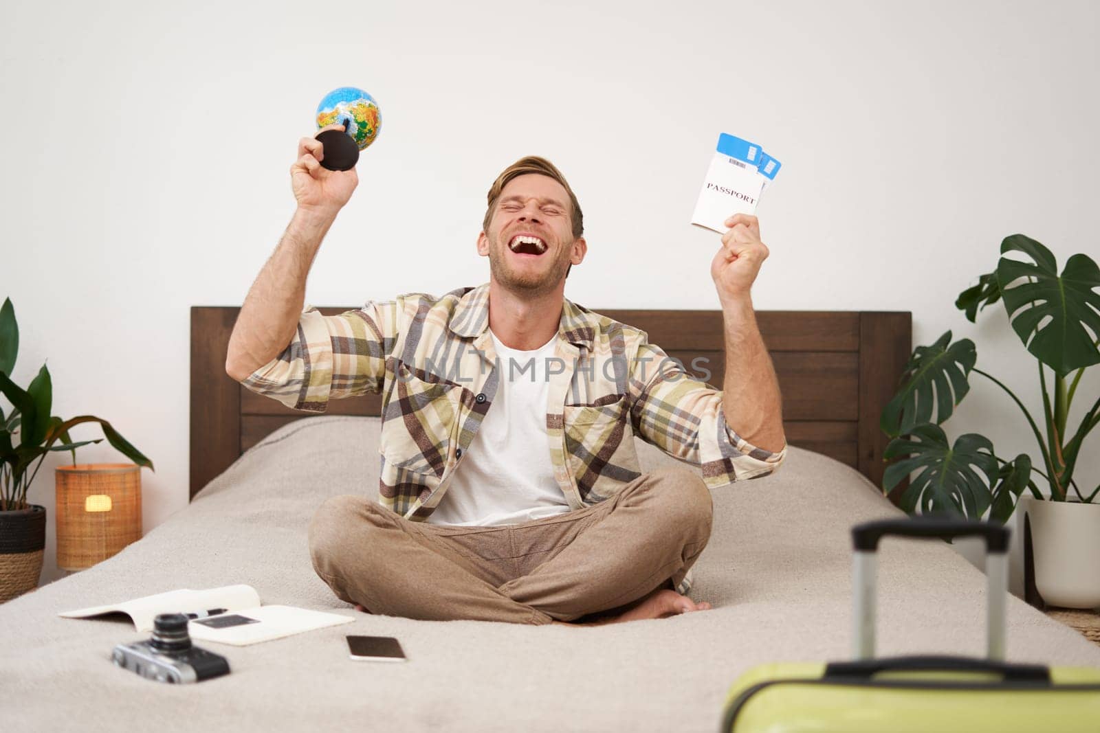 Portrait of happy guy, tourist goes on vacation abroad, holds globe and plane ticket, excited about his holiday, sits on bed with smiling, enthusiastic face expression. Copy space