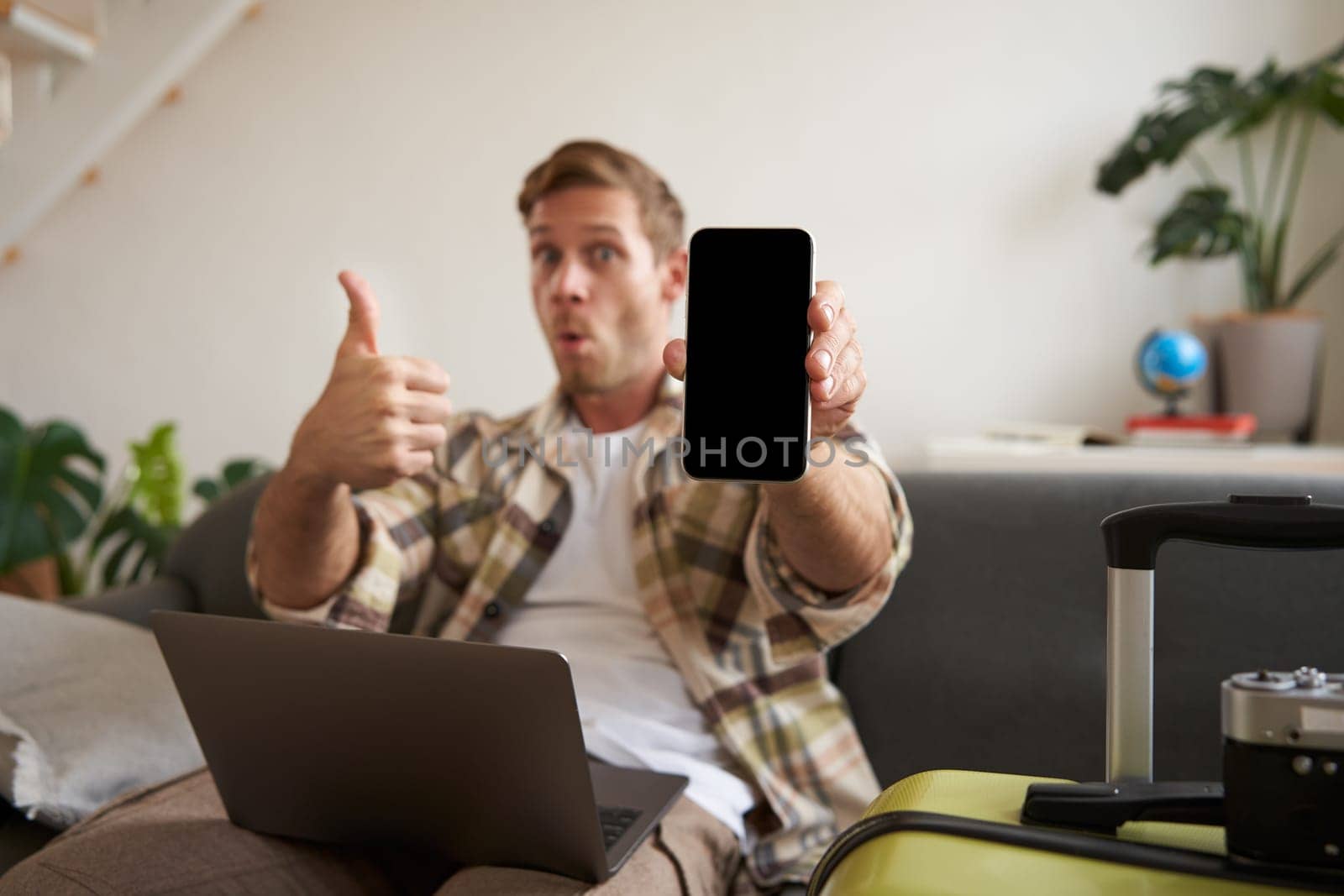 Portrait of handsome young man, showing smartphone screen, recommending mobile app for travelling or booking online, sitting with laptop and suitcase by Benzoix