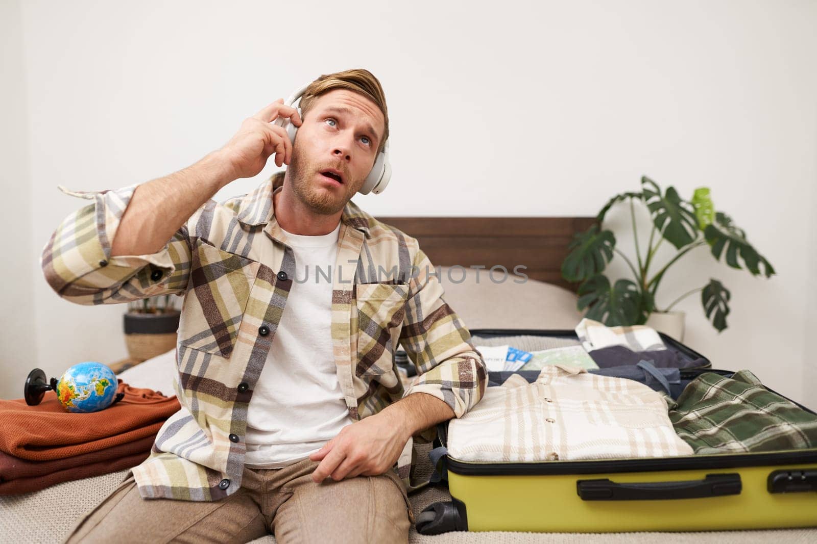 Portrait of handsome young man packing things in suitcase, listening to music in headphones, tourist going on vacation, preparing for holiday trip.