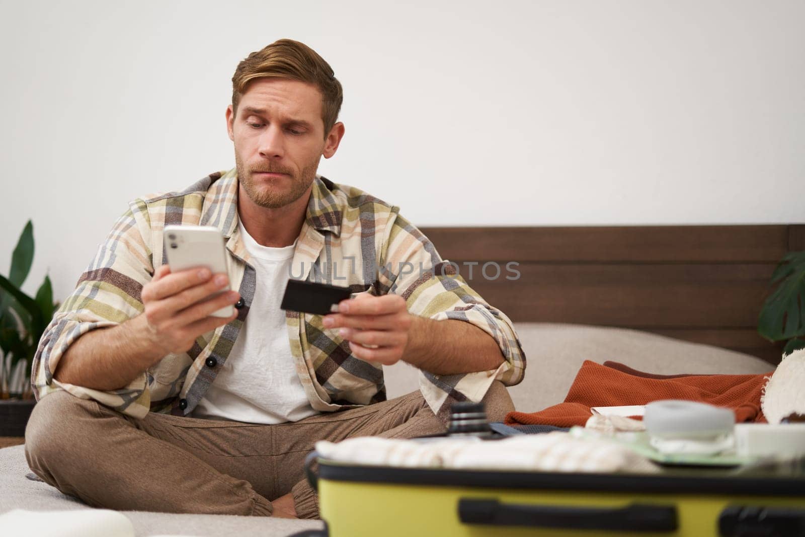 Angry man holds credit card and mobile phone, sits next to clothes in suitcase, has problems with paying online.