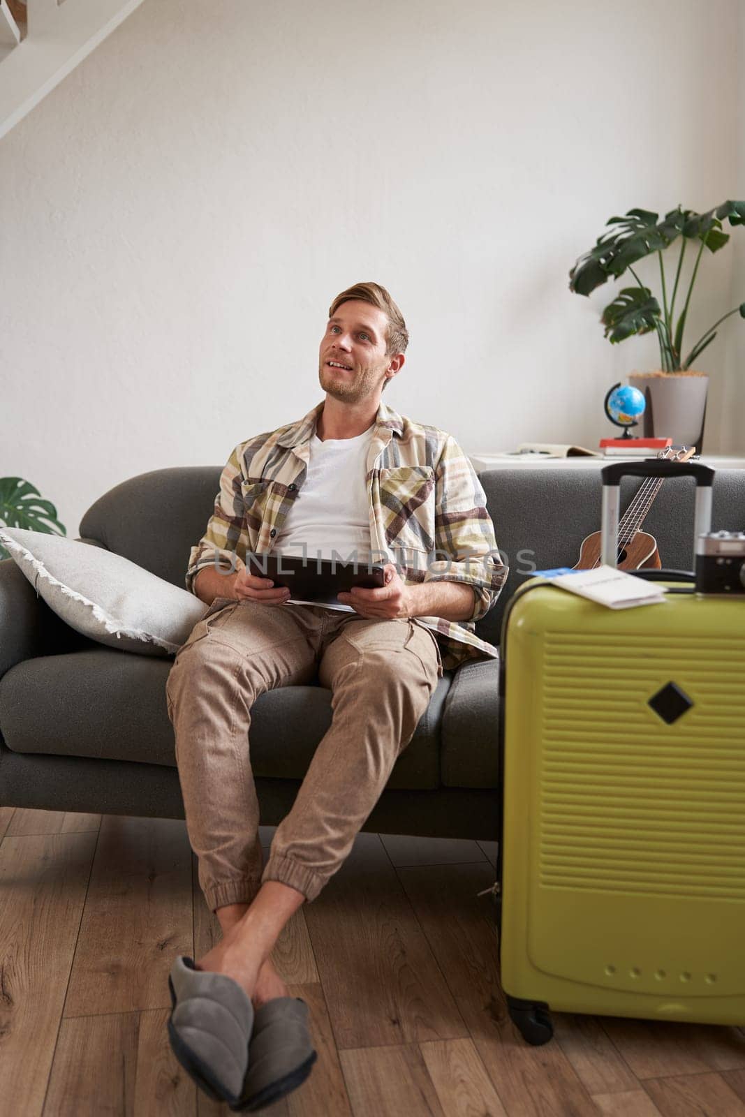 Image of young man, tourist goes on holiday, packed suitcase, sits with digital tablet on sofa, reads, looks at his map, books hotel for business trip by Benzoix