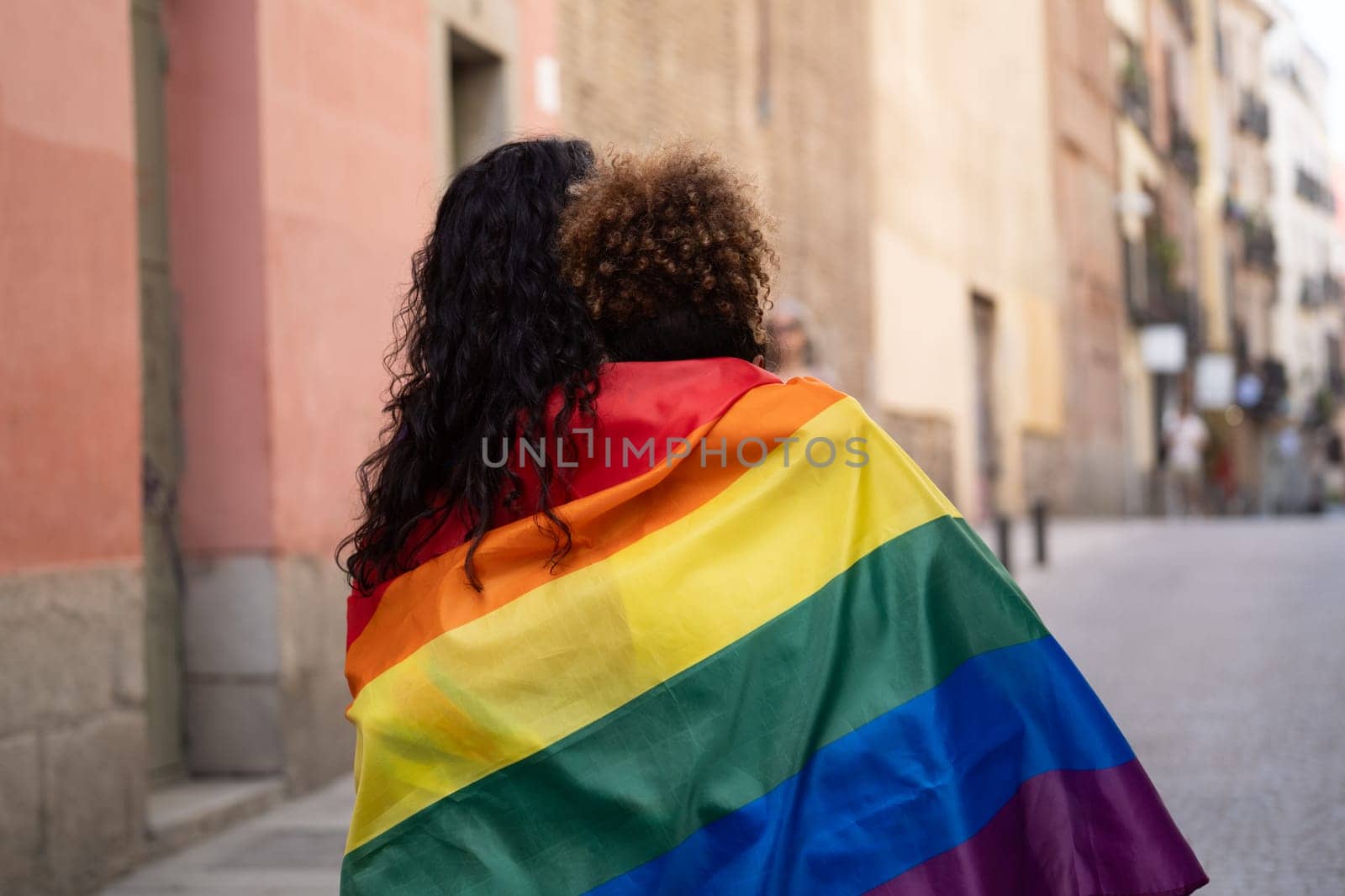 Rear view of a couple of women covered with rainbow flag in a urban street. Lgbt love pride concept.
