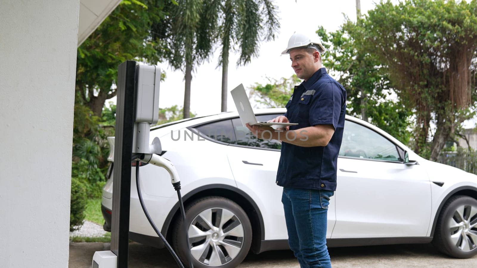 Qualified technician working on home EV charging station installation, making troubleshooting and configuration setup on charging system with laptop for EV at home. Synchronos