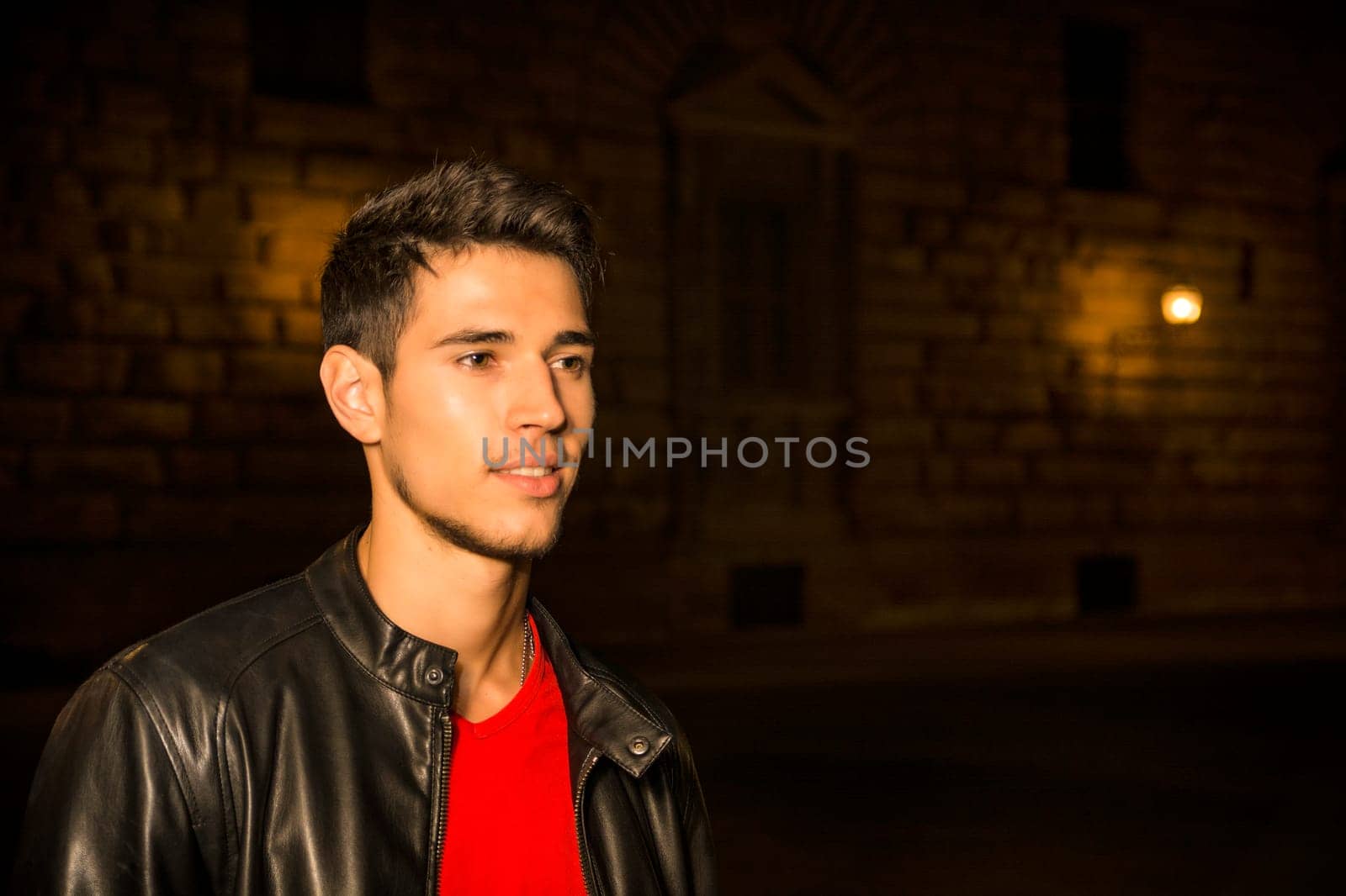 A man in a black leather jacket standing in a dark city street at night