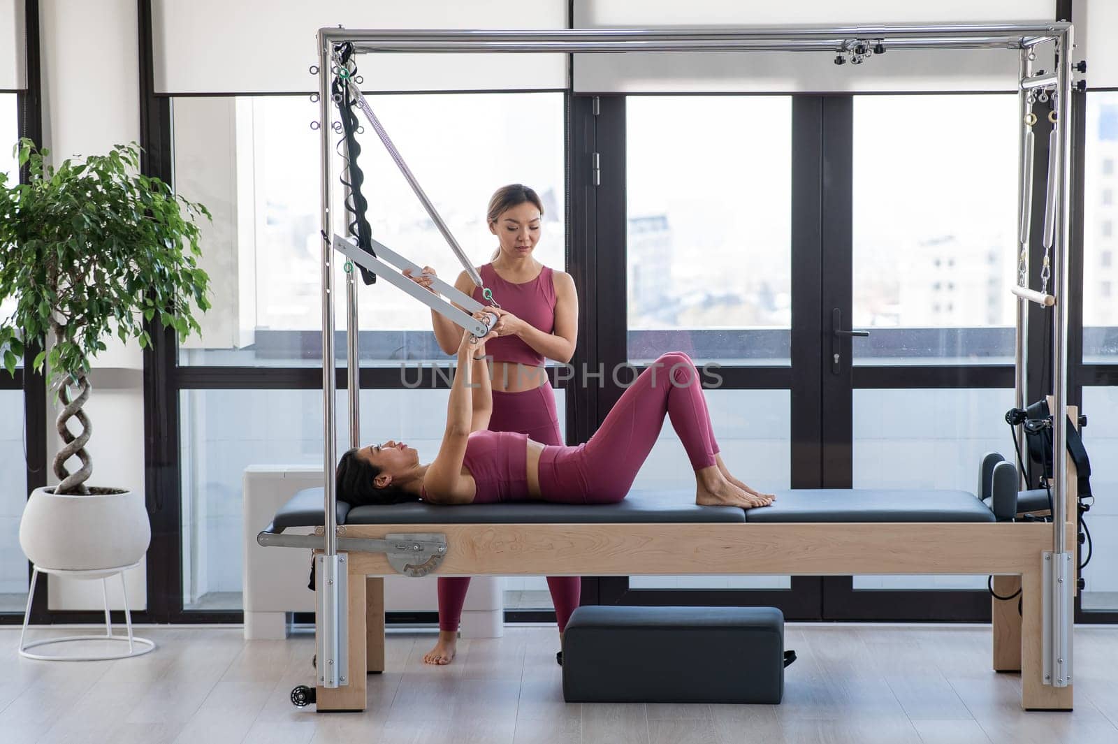 Asian woman doing pilates with trainer on cadillac reformer