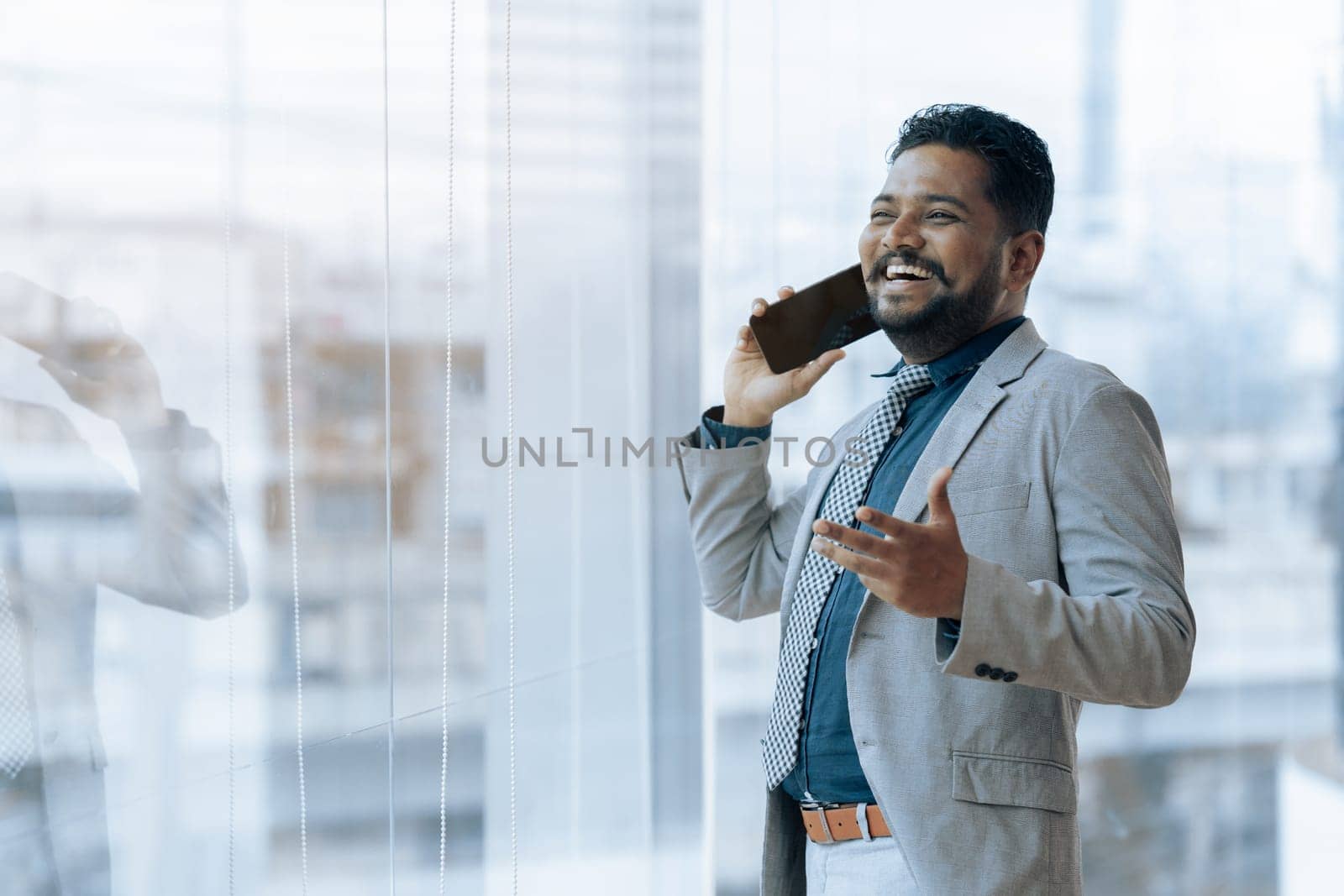 Indian businessman using smartphone talking having phone call conversation in office looking out window.