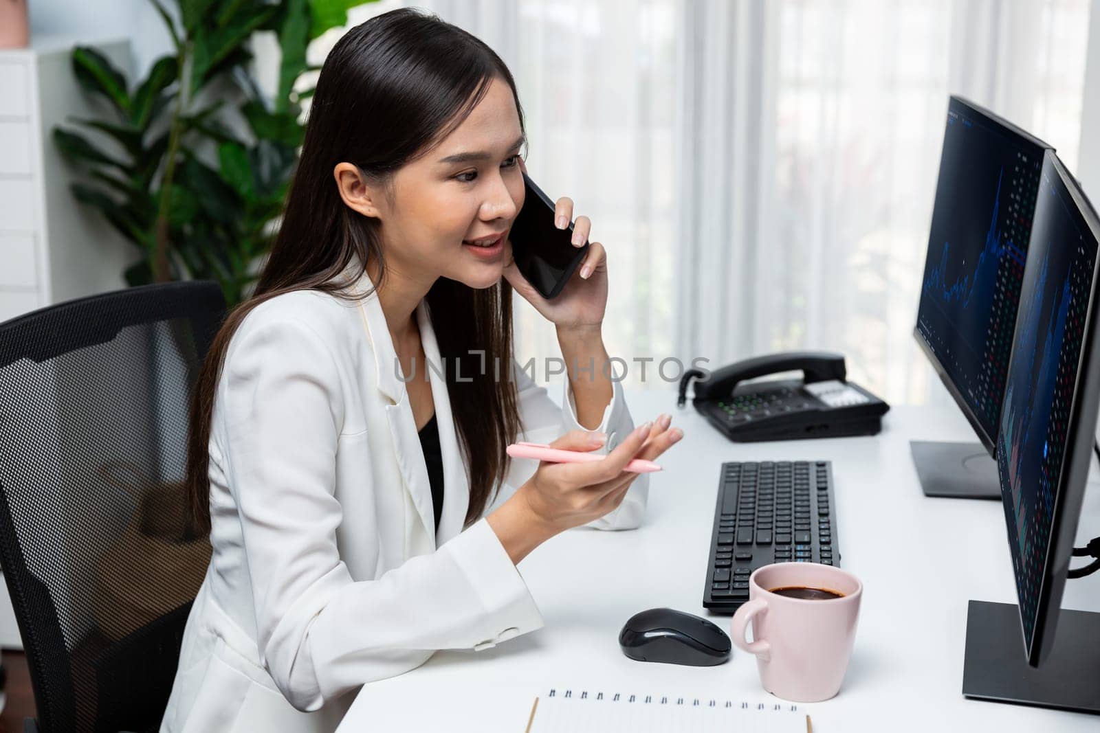 Smiling young beautiful Asian businesswoman calling to broker on smartphone to analyze exchange stock market graph on pc screen at modern office. Concept of investing high profit in wealth. Stratagem.