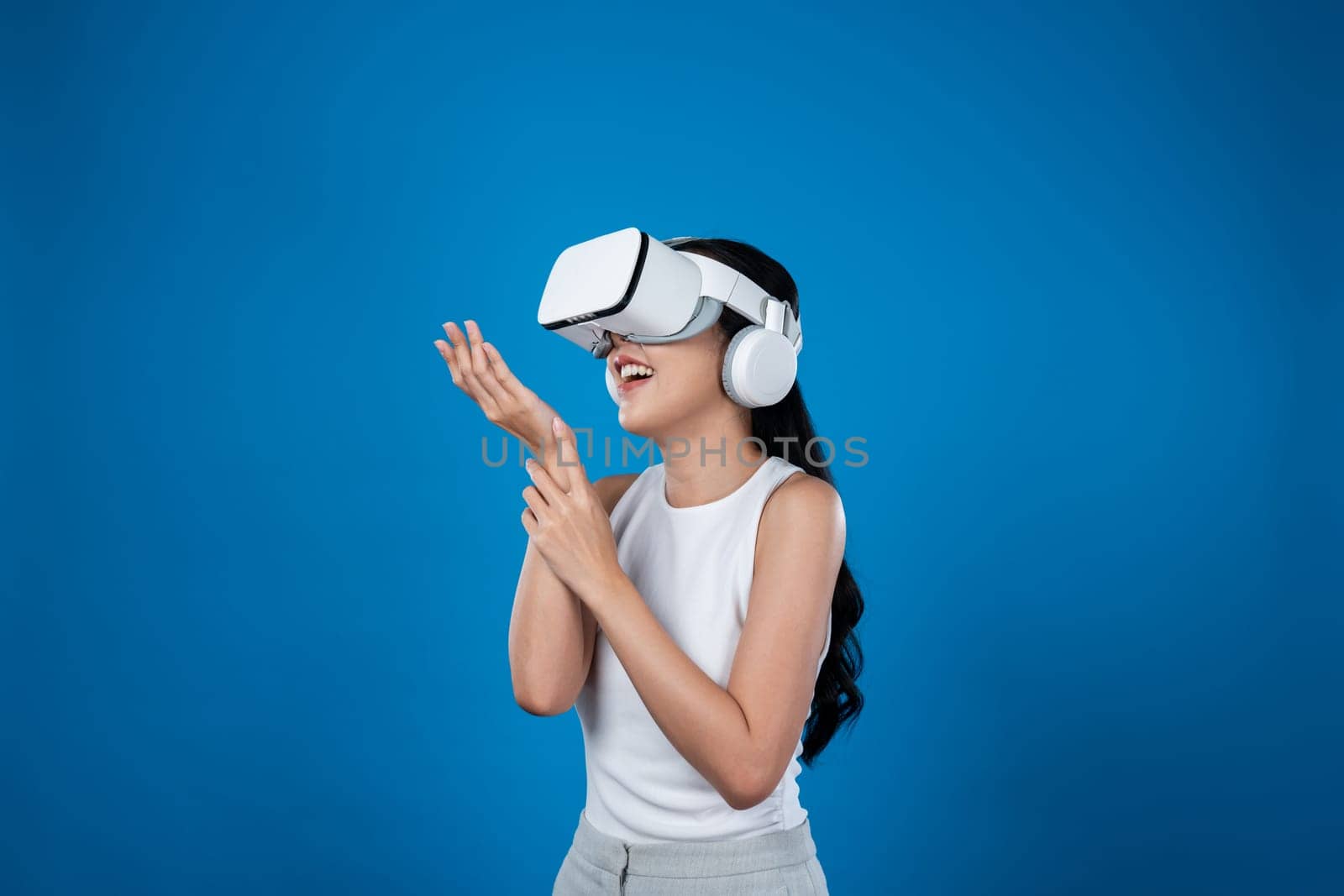 Smart female standing with blue background wearing VR headset connecting metaverse, futuristic cyberspace community technology. Elegant woman excited seeing generated virtual scenery. Hallucination.