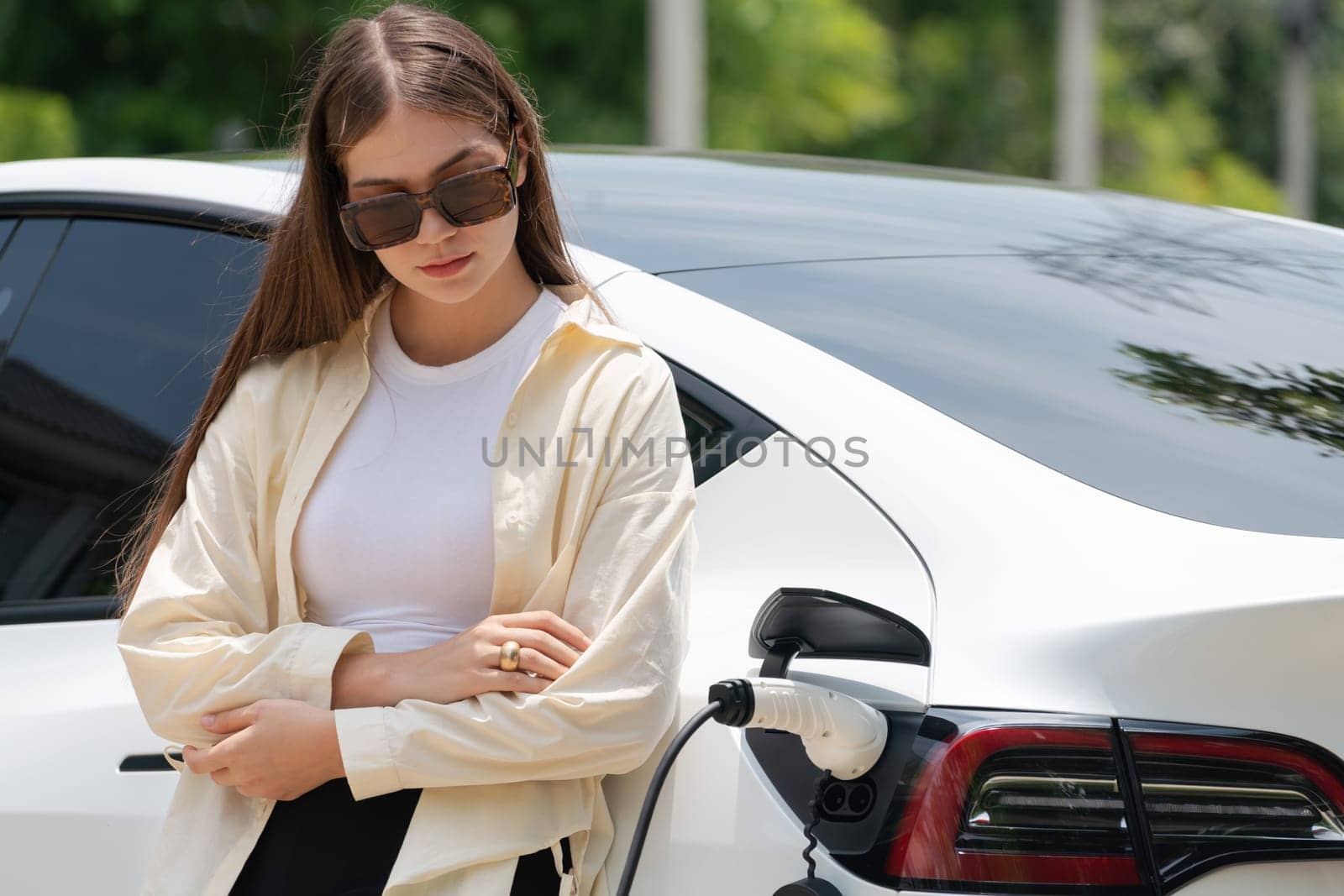 Young woman recharge her EV electric vehicle at green city park parking lot. Urban sustainability lifestyle for environmental friendly EV car with battery charging station. Expedient