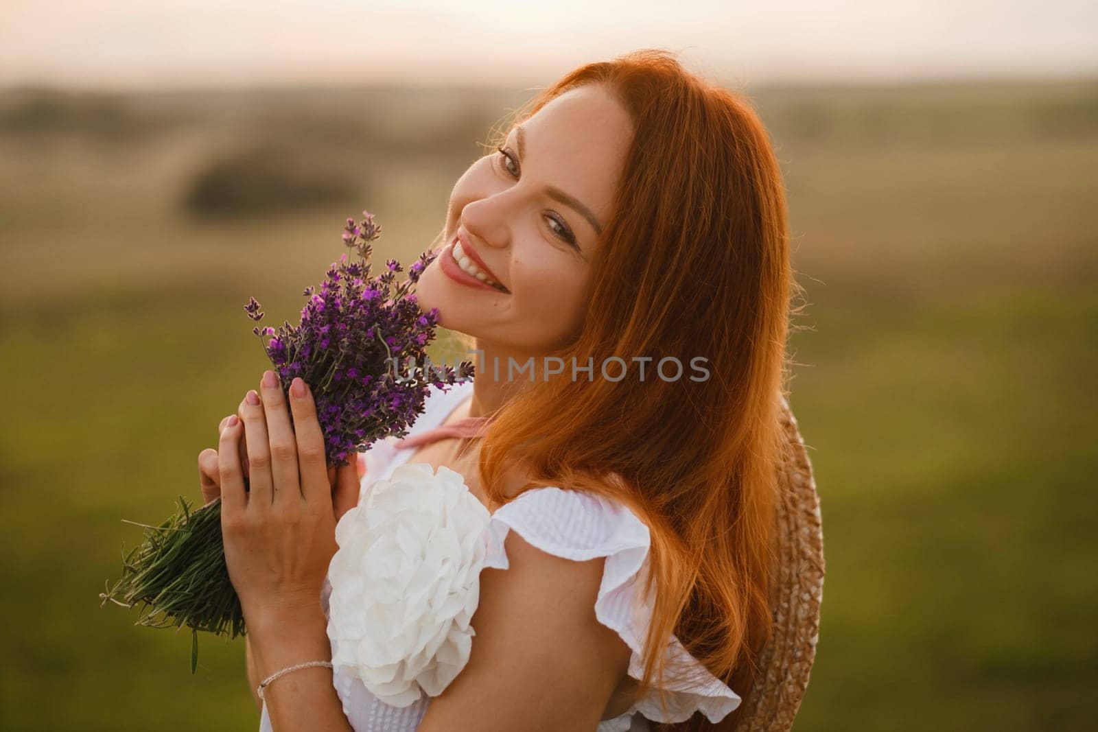 A girl in a white dress with a bouquet of lavender flowers stands in a field at sunset.