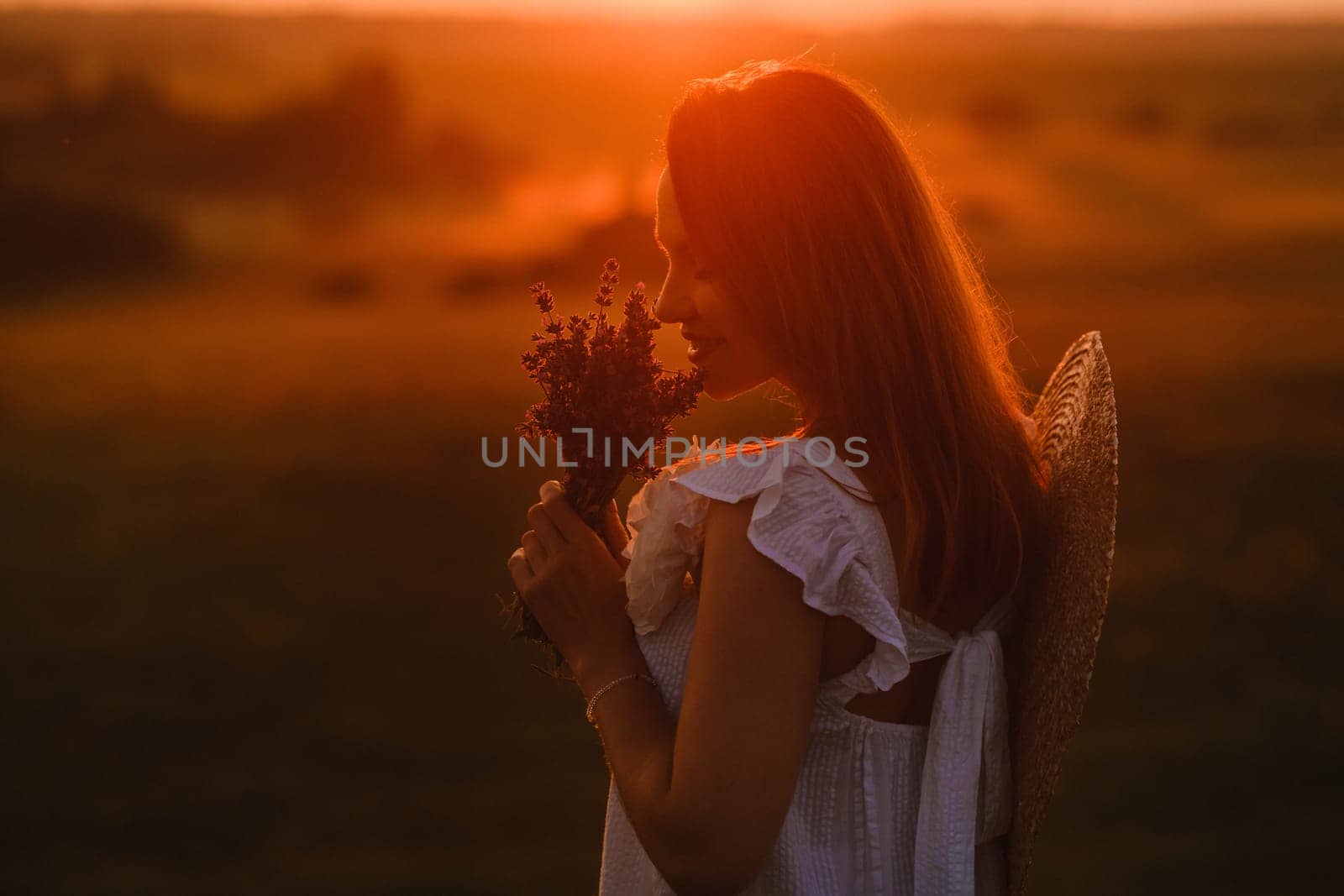 A girl in a white dress with a bouquet of lavender flowers stands in a field at sunset by Lobachad