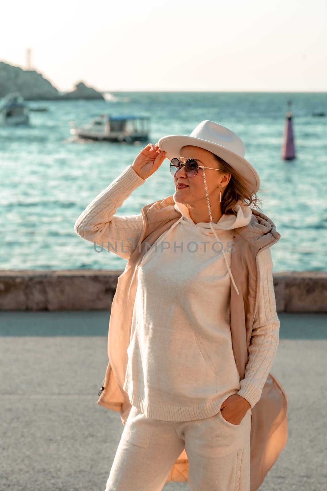 Happy blonde woman in a white suit and hat posing at the camera against the backdrop of the sea.