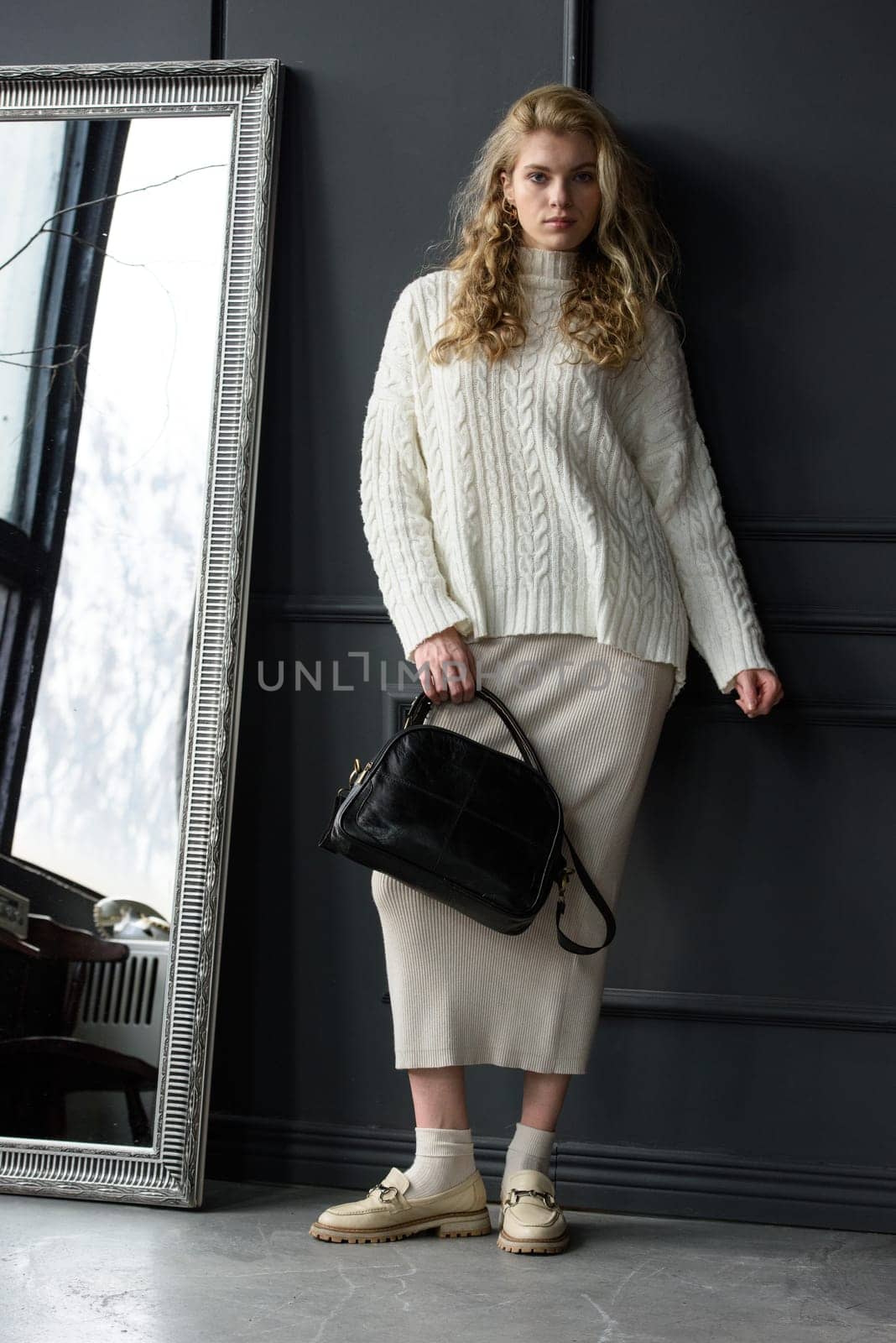 Studio portrait of beautiful woman with a curly blond hair holding black bag, posing on gray background. Model wearing stylish sweater and skirt