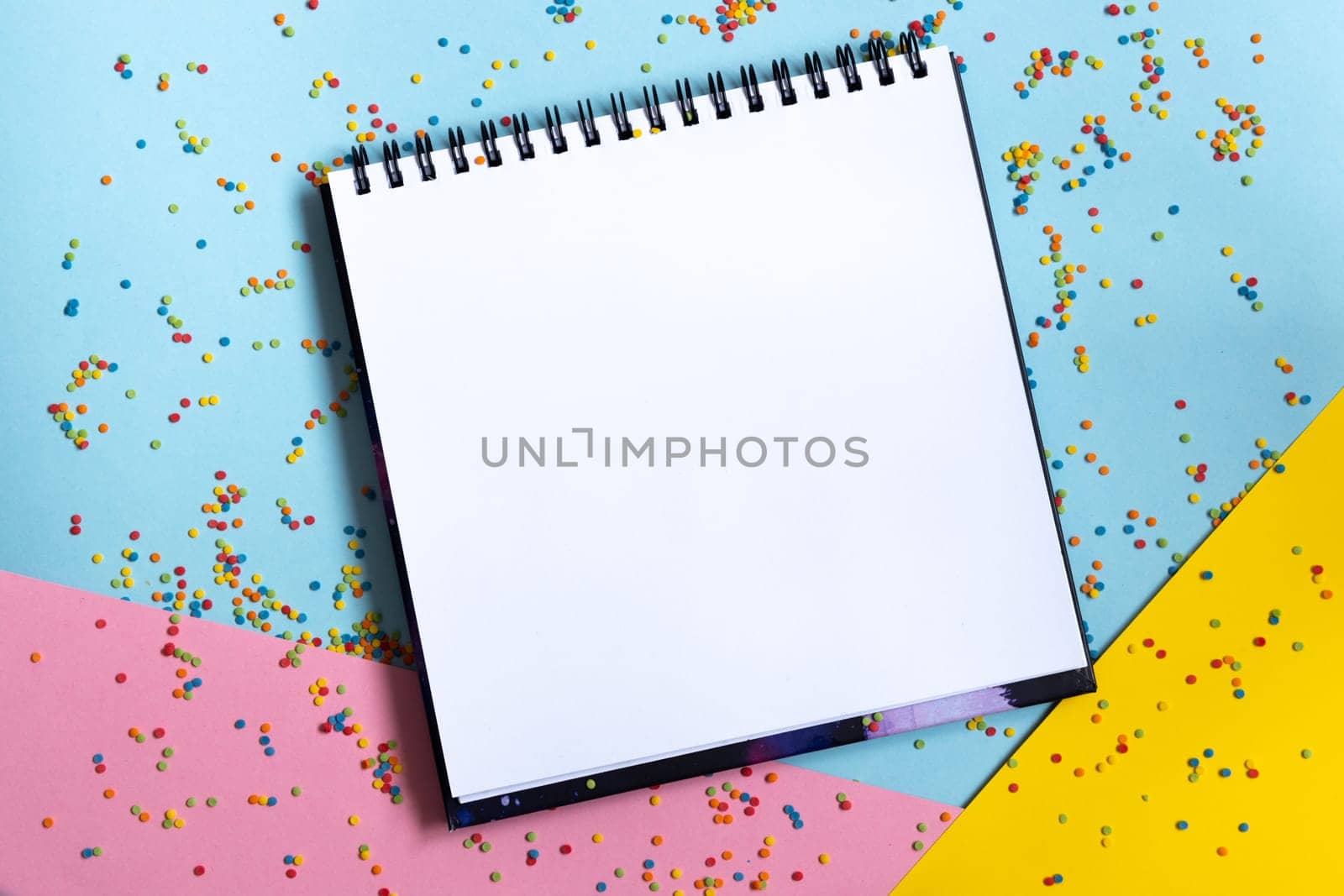 Mock up of empty spiral scketchbook with white paper on pastel blue background with colorful confetti . Top view of open notebook with clean sheet. Template for message. Copy space