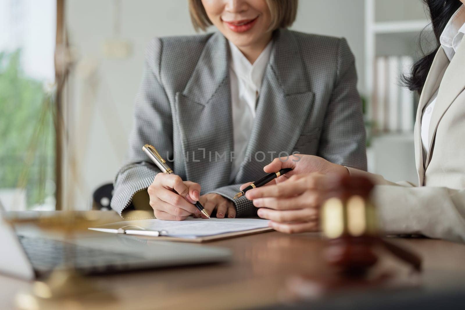 A woman is signing a document at a desk with a laptop and a chair. The woman is wearing a suit and is writing with a pen. The scene suggests a professional setting