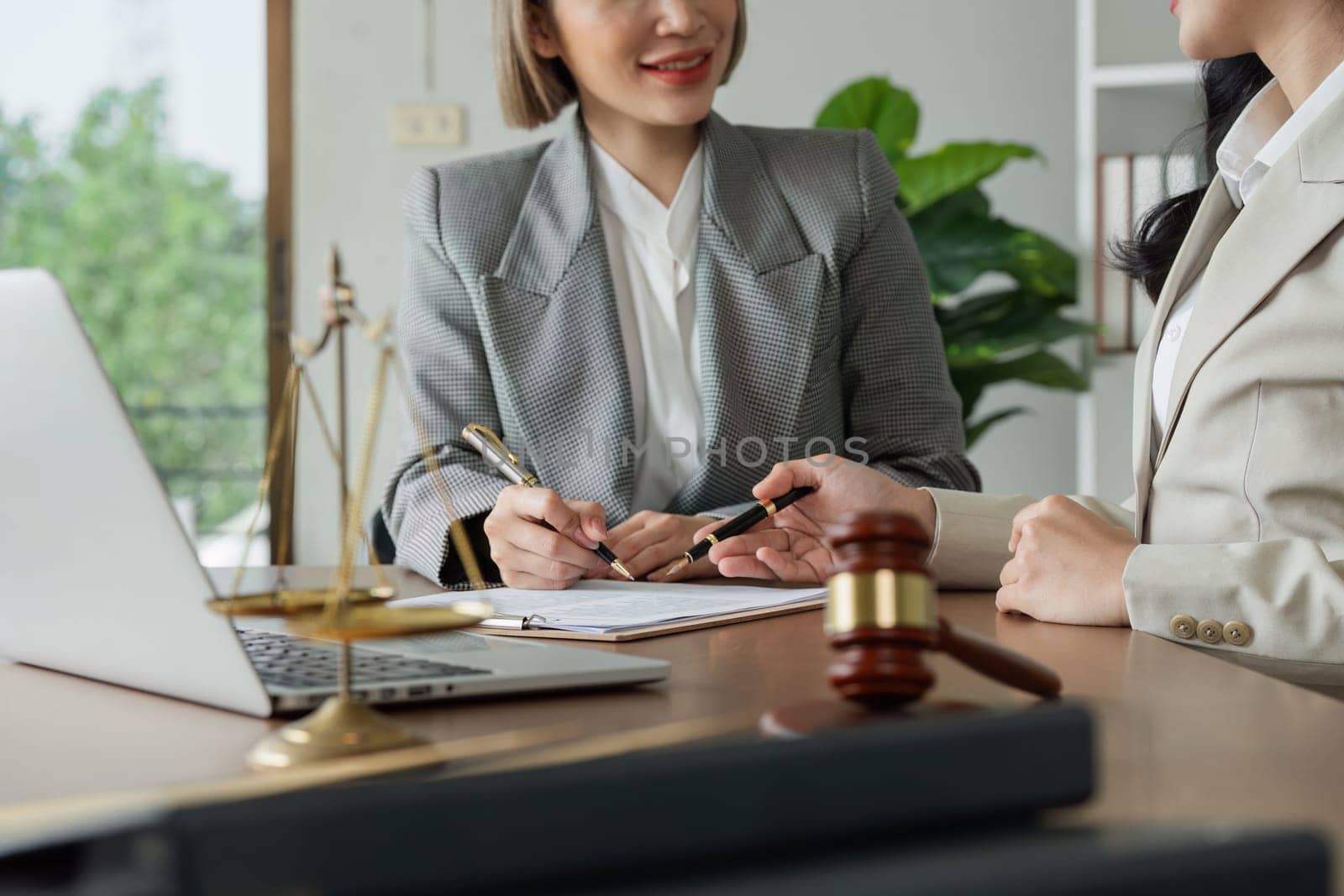 A woman is signing a document at a desk with a laptop and a chair. The woman is wearing a suit and is writing with a pen. The scene suggests a professional setting