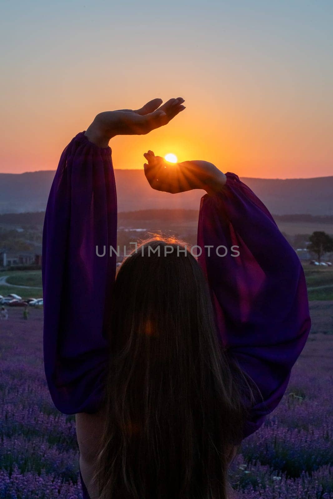 Woman raises arms in lavander field at sunset enjoys sunset in purple flower field. Serene floral setting. Setting sun. Conveying peaceful ambiance in flower field at sunset
