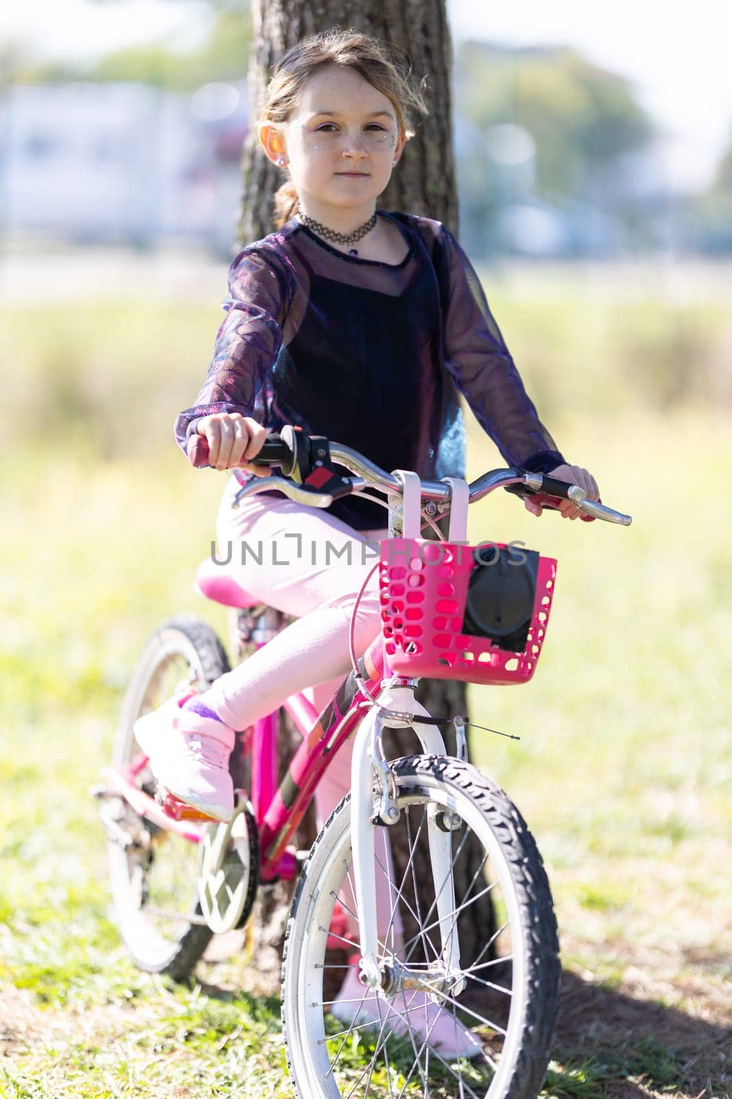 A young girl is sitting on a pink bicycle with a basket. She is wearing a black shirt and pink pants