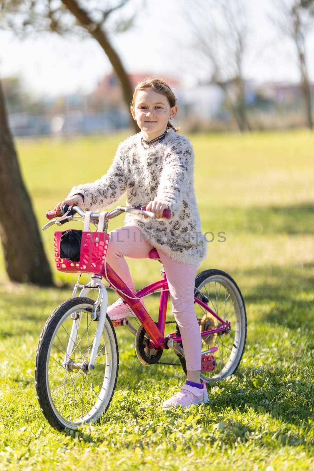 A young girl is sitting on a pink bicycle in a park. She is wearing a pink sweater and pink pants