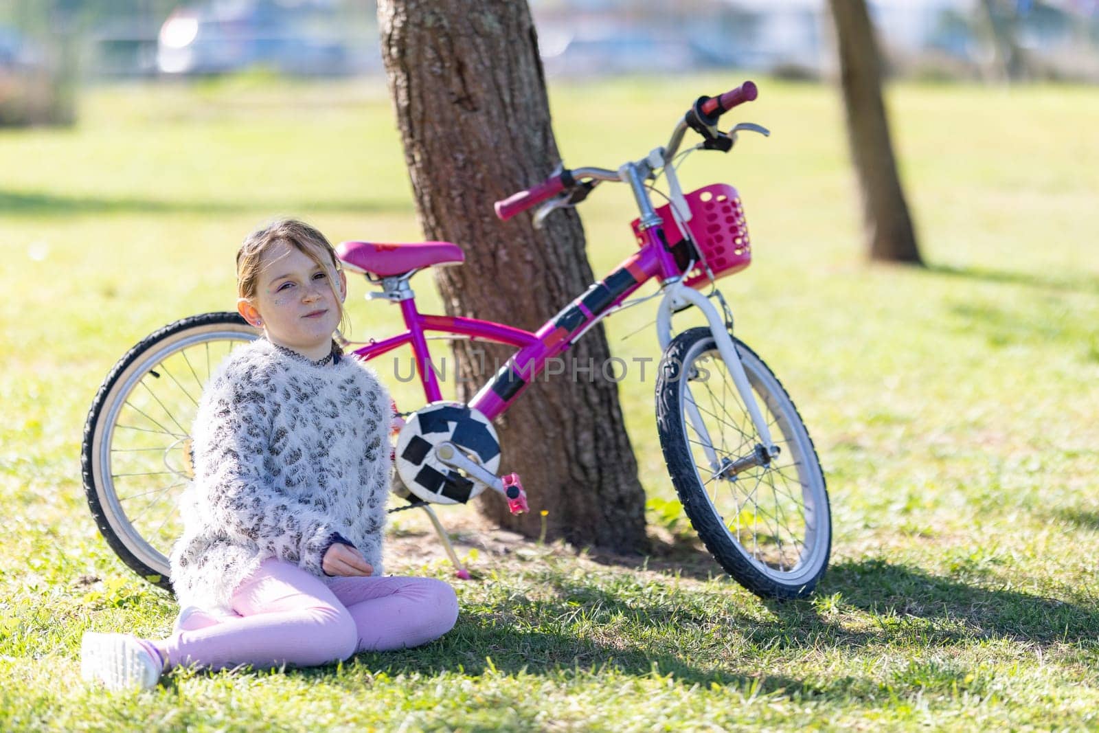 A young girl sits on the grass next to a pink bicycle. The girl is wearing a white sweater and pink pants