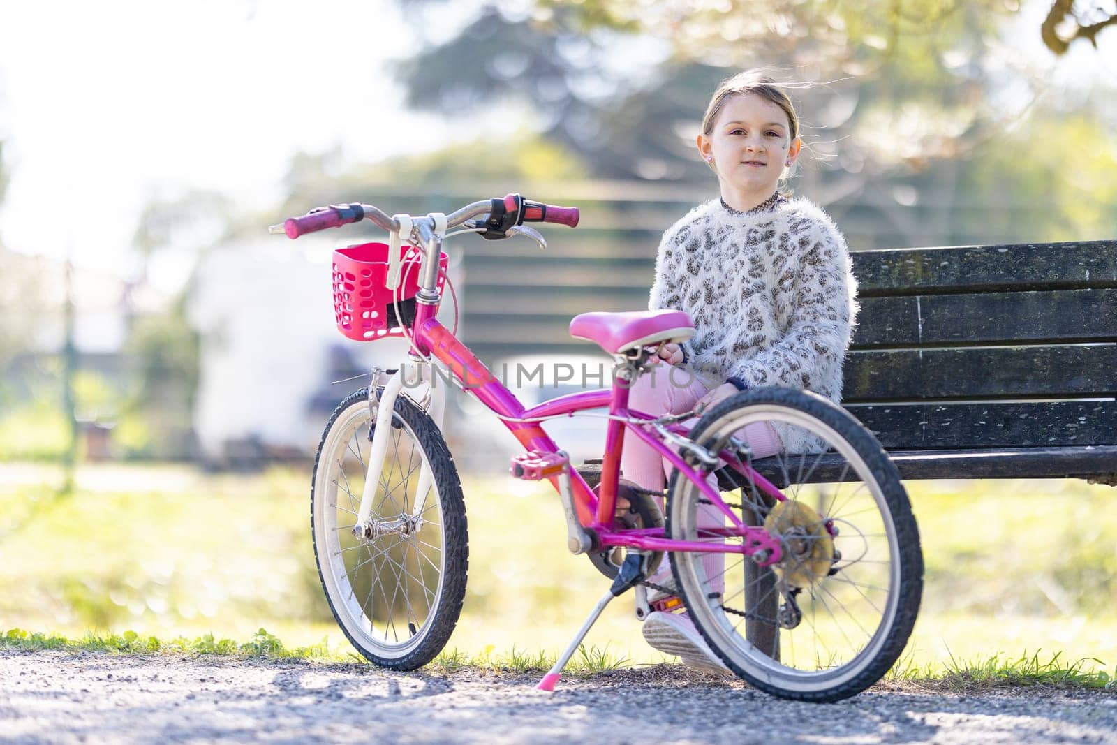 A young girl sits on a bench next to a pink bicycle. The bicycle has a basket on the front and a pink seat. The girl is smiling and he is enjoying her time outdoors