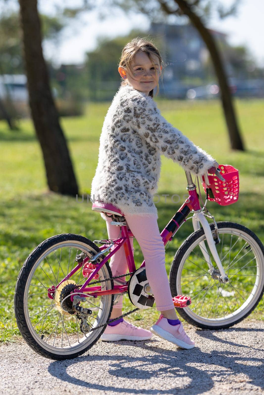 A young girl is sitting on a pink bicycle with a basket. She is wearing a white sweater and pink pants