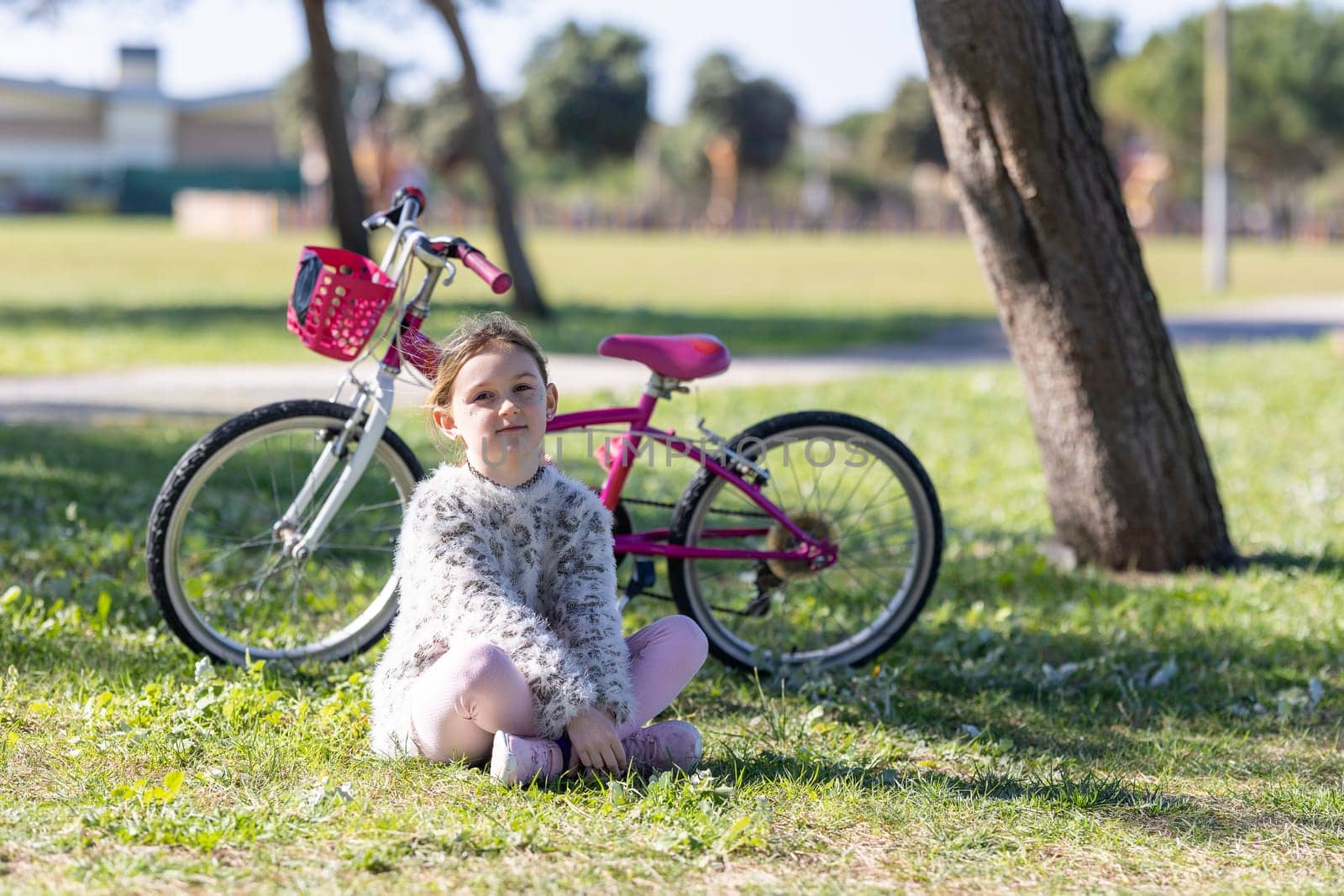 A little girl is sitting on the grass next to a bicycle. She is wearing a pink shirt and white pants