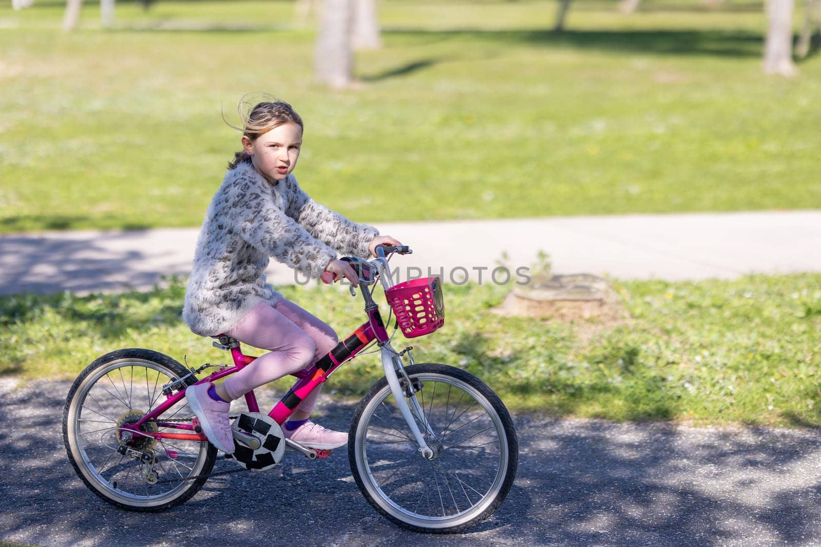 A young girl is riding a pink bicycle with a basket on the front. She is wearing a pink sweater and pink pants