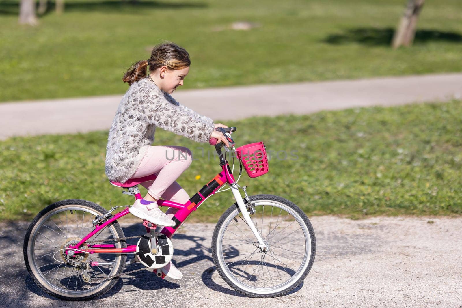 A young girl is riding a pink bike with a basket on the front. She is wearing a pink jacket and pink pants. The scene is set in a park with a grassy area