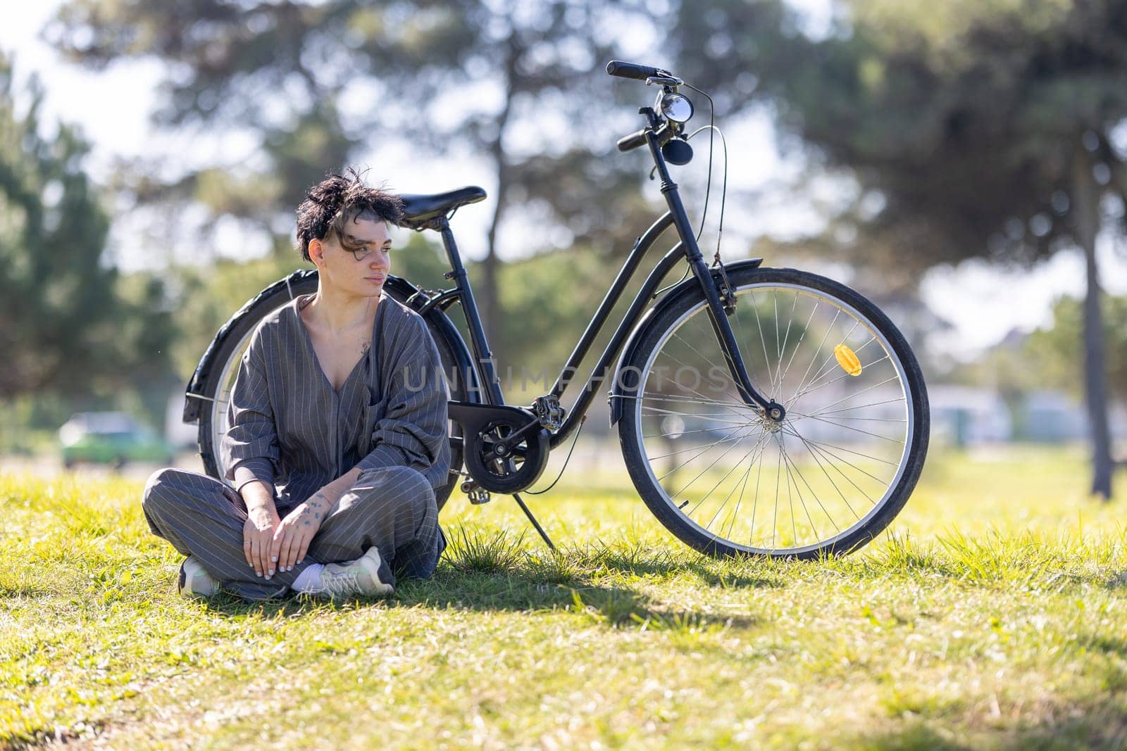 A woman sits on the grass next to a black bicycle by Studia72