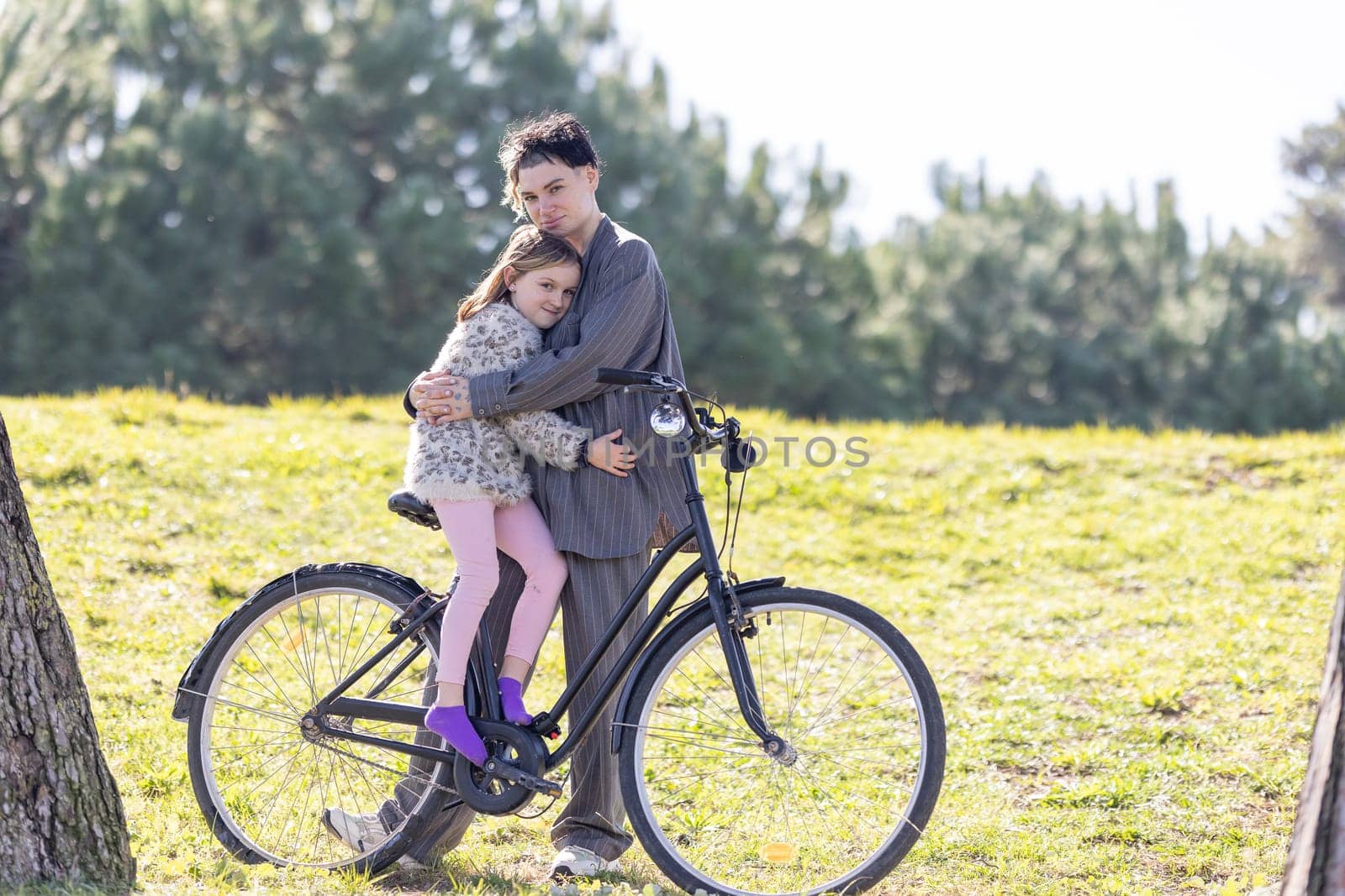 A woman and a little girl - her daughter - are hugging each other while sitting on a bicycle. The scene is peaceful and heartwarming, with the two of them enjoying each other's company in a beautiful outdoor setting