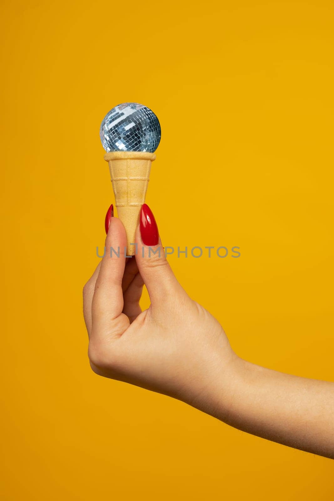 Woman hand holding small crispy ice cream cone with a mirrored disco ball isolated on yellow background by zartarn
