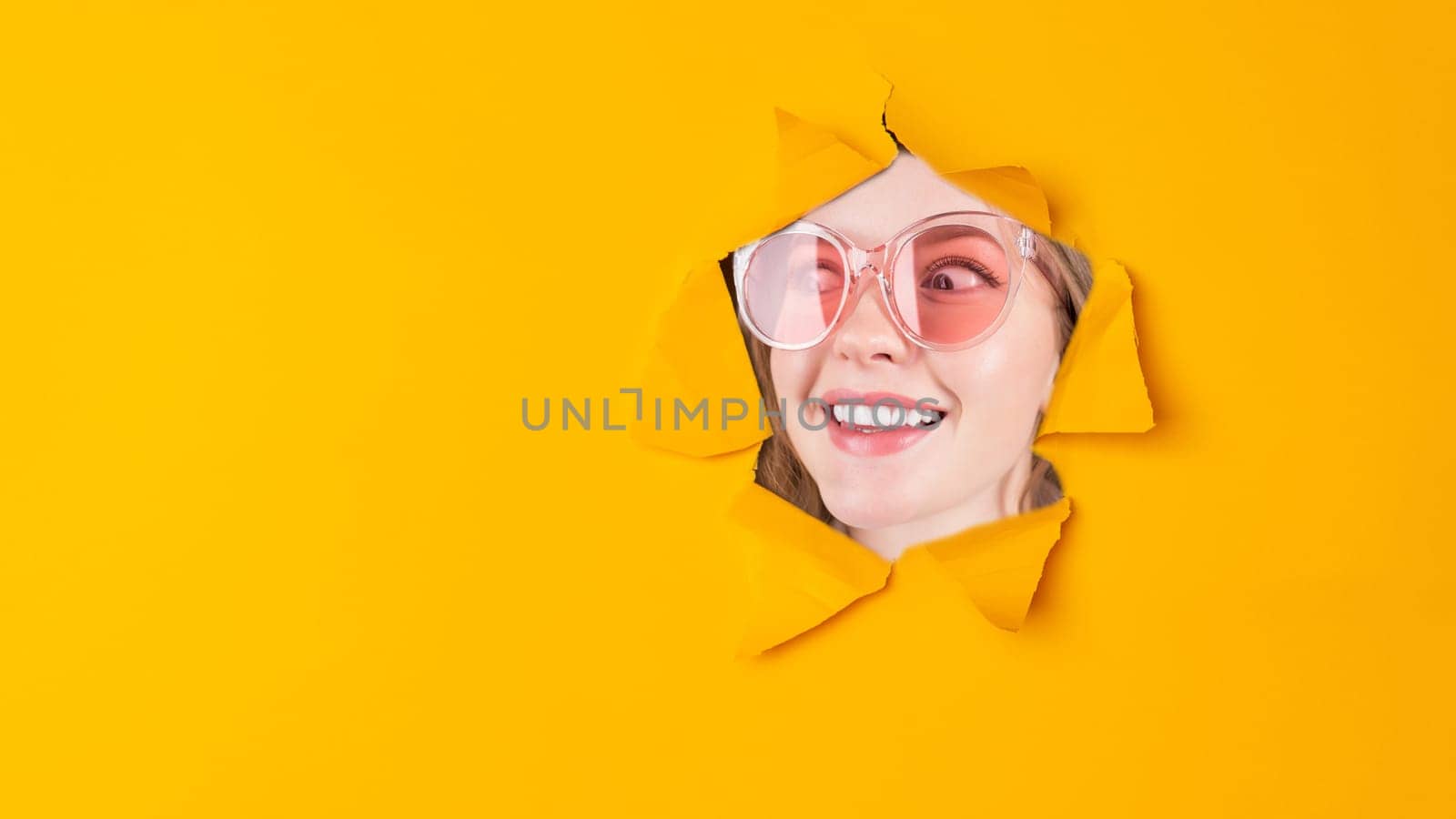Beautiful face of young smiling woman with perfect teeth looking in paper torn hole in yellow background with copy space.