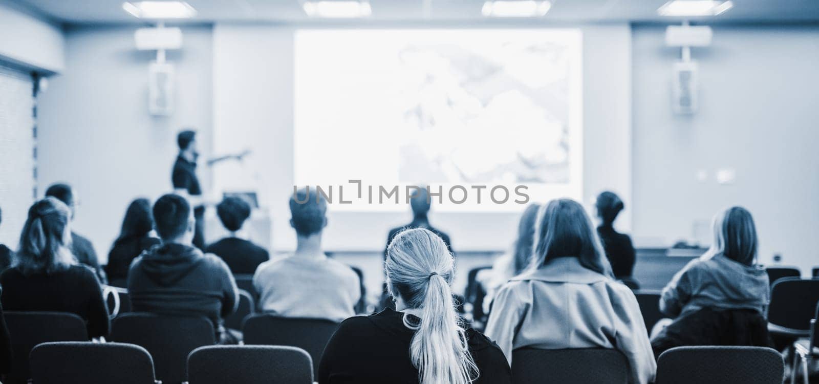 Speaker giving a talk in conference hall at business event. Rear view of unrecognizable people in audience at the conference hall. Business and entrepreneurship concept. Blue tones black and white