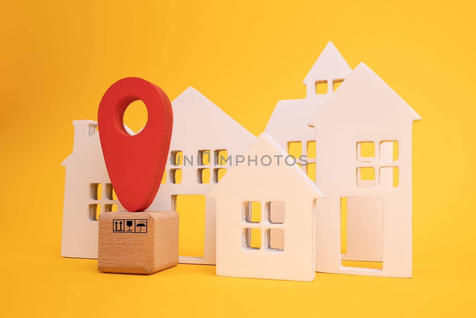 Cardboard boxes and a red position pin in city of white toy house on yellow background. Locating packages and goods.