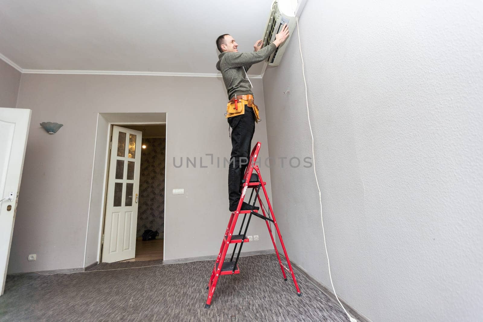 Professional electrician man with screwdriver maintaining, cleaning modern air conditioner indoors. Young technician standing on a ladder in the room repairing or installing air conditioner. High quality photo