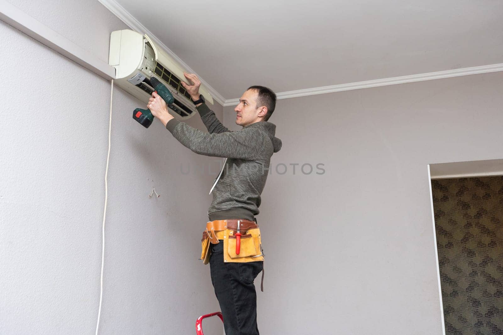 Professional electrician man with screwdriver maintaining, cleaning modern air conditioner indoors. Young technician standing on a ladder in the room repairing or installing air conditioner. High quality photo