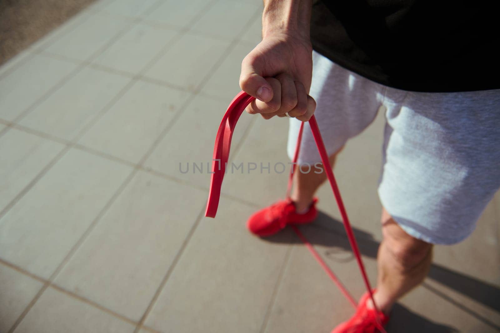 Top view of sportsman in red sneakers, gray sports shorts and black t-shirt doing warm up exercises with resistance band by artgf